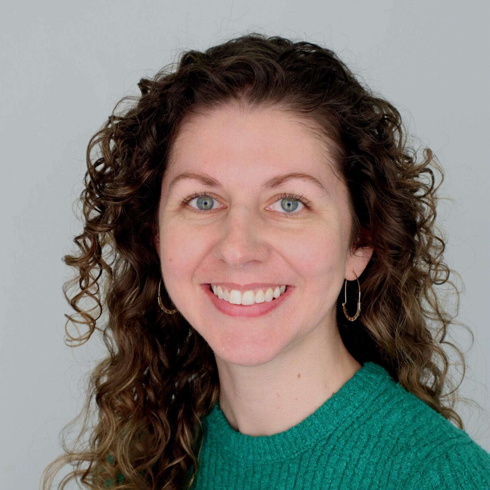 Woman with curly brown hair and blue eyes, wearing a green sweater and gold hoop earrings, smiling at the camera against a plain light background.