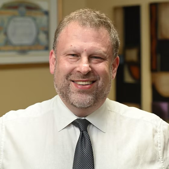 A middle-aged man with short curly hair and a beard smiles at the camera, wearing a white dress shirt and dark tie, with abstract art in the background.