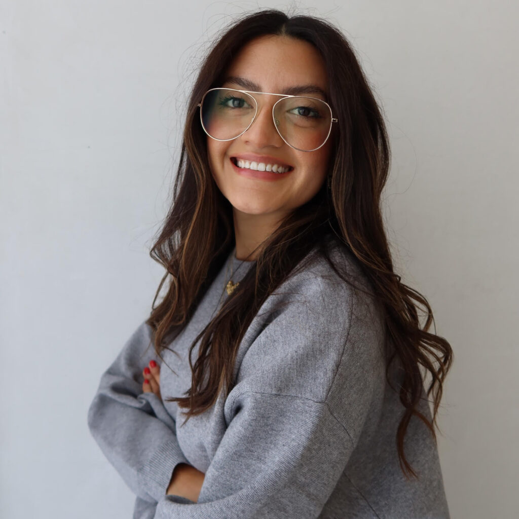 Woman with long brown hair and large aviator glasses smiles at the camera, wearing a grey sweater and standing against a plain white background.