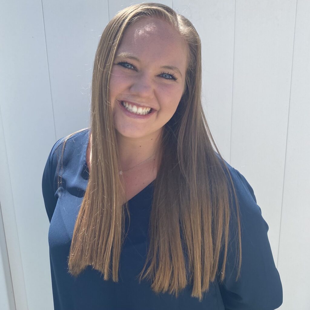 A woman with long straight hair wearing a navy blue top smiles at the camera while standing in front of a white background.