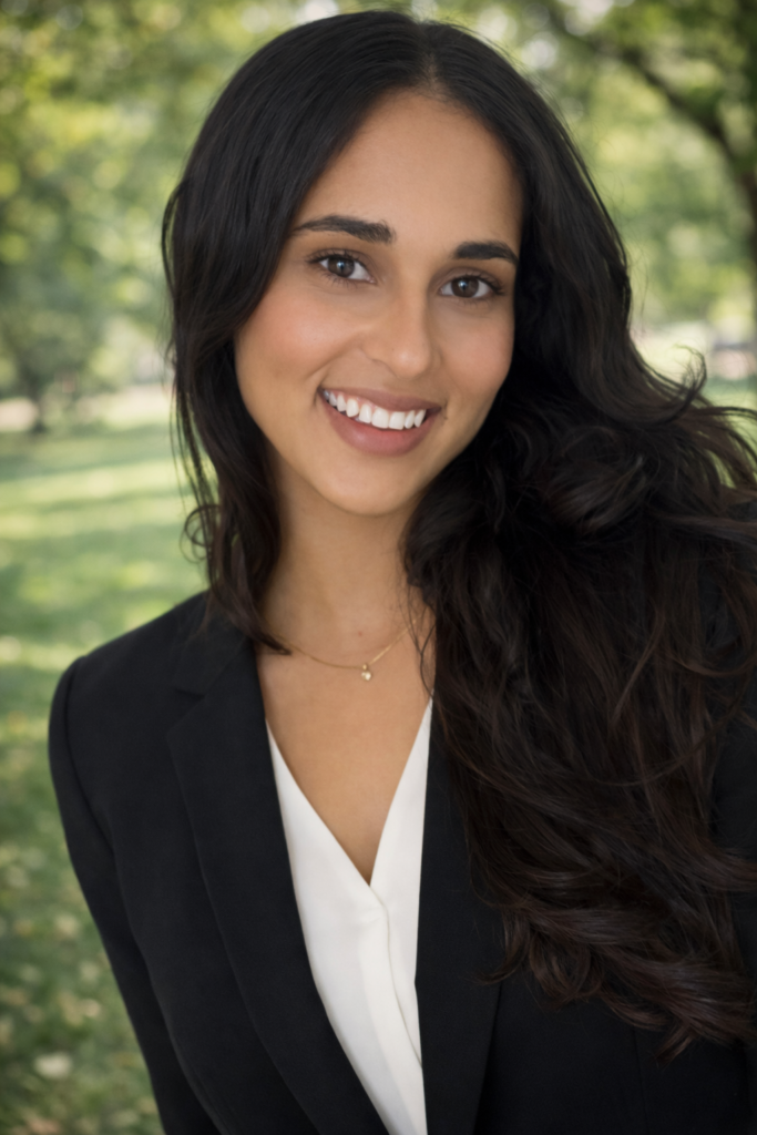 A woman with long dark hair and a black blazer smiles outdoors with green trees in the background.