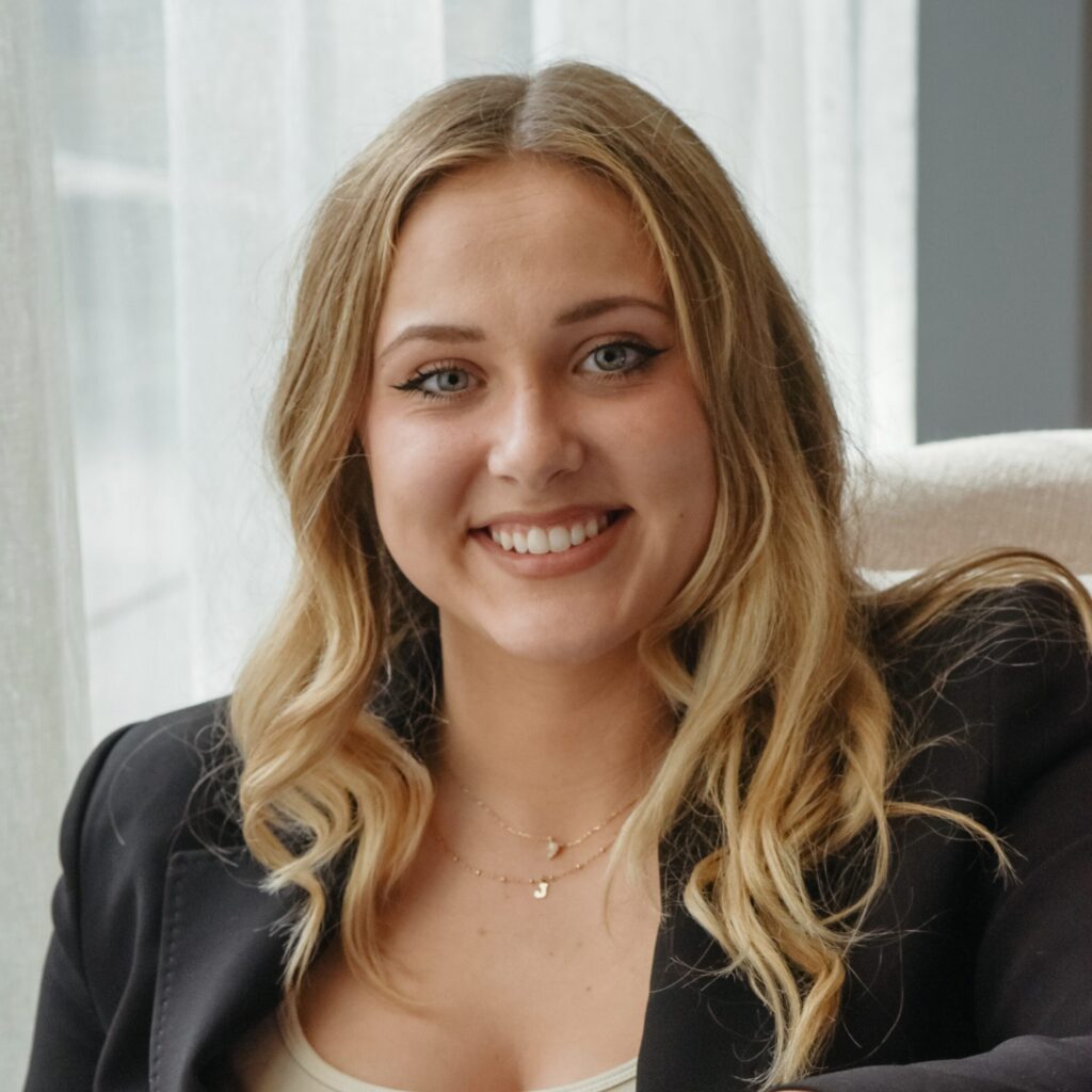 A woman with long blonde hair, wearing a black blazer and light top, sits indoors on a cream chair, smiling at the camera.
