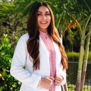 A woman with long brown hair, wearing a white lab coat over a pink dress, stands outdoors in front of palm trees and greenery, smiling at the camera.