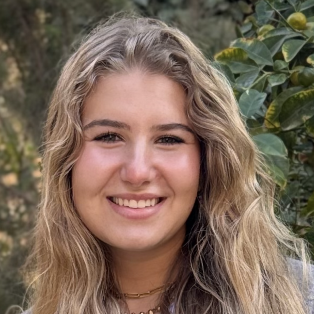 A young woman with long wavy blonde hair, wearing a grey coat over a blue and white striped shirt, smiles while standing outdoors in front of greenery.