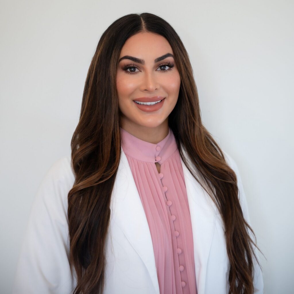 A woman with long brown hair wearing a white coat over a pink blouse stands in front of a plain light background, smiling at the camera.