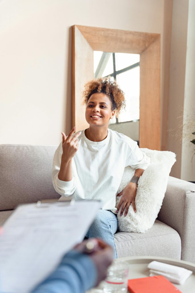 A woman sits on a sofa, smiling and gesturing while talking to someone holding a clipboard in the foreground.