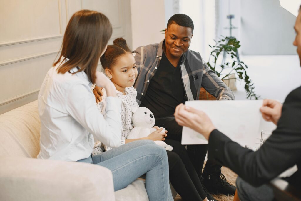 A family with a young girl holding a stuffed animal sits on a couch, talking to a person holding a clipboard in a bright, modern room.