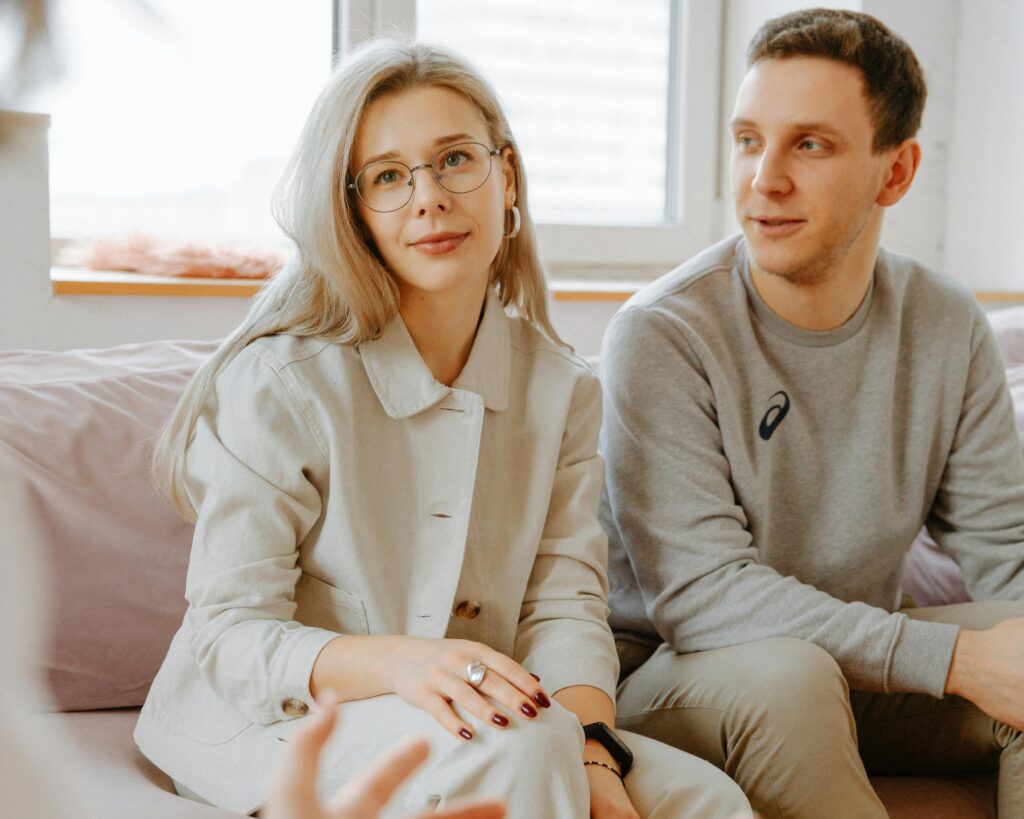 A woman with glasses and a man sit on a couch, listening to someone out of frame. The setting appears to be a casual indoor environment.