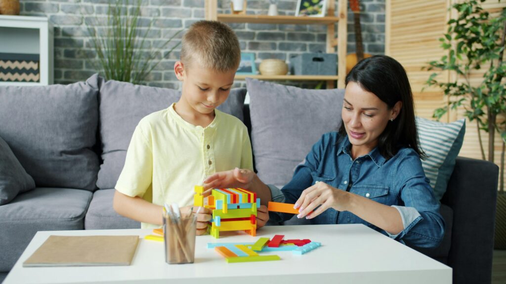 A woman and a child sit at a white table, building a structure with colorful blocks in a living room setting.