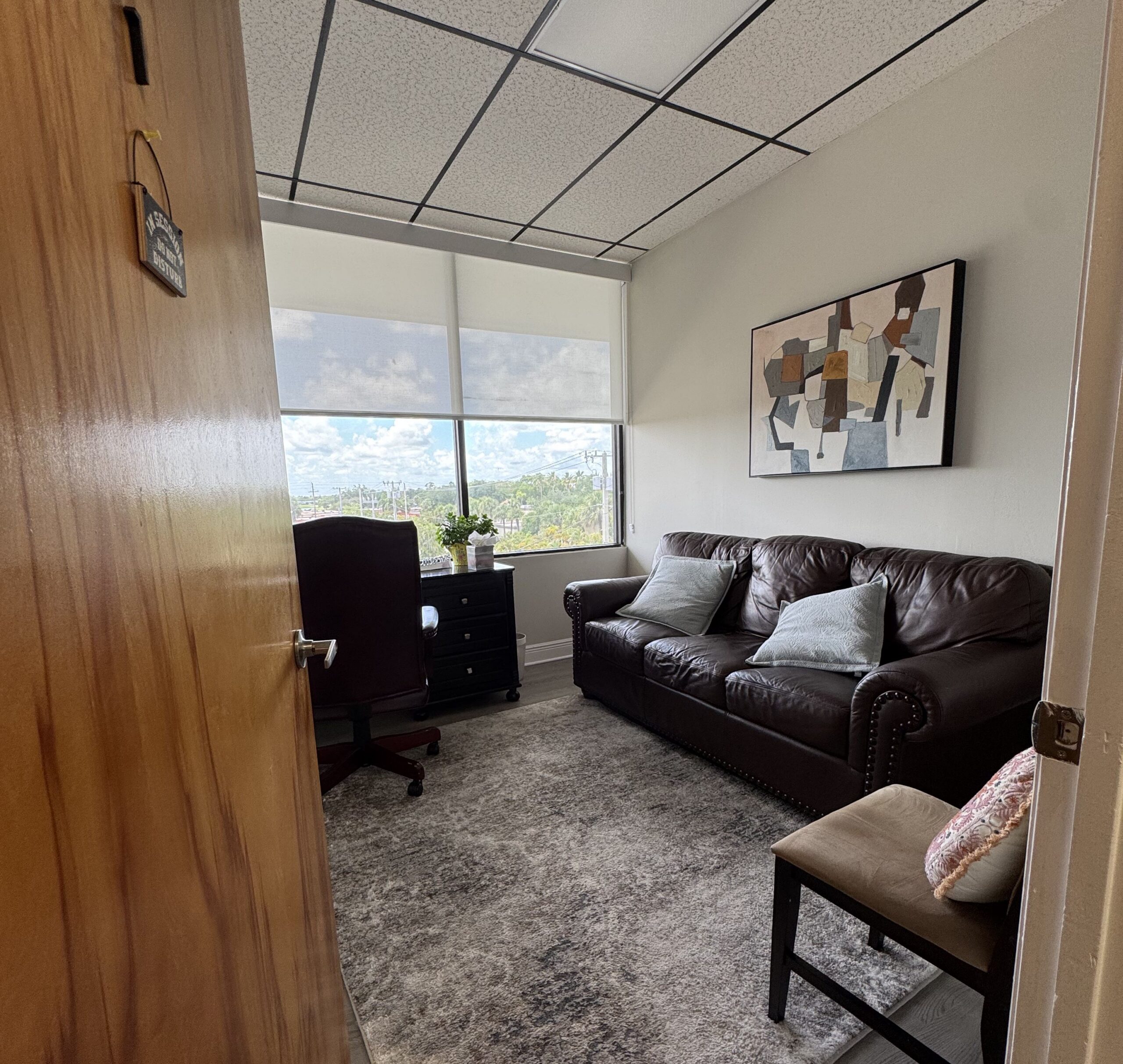 A small office with a leather sofa, cushioned chair, desk, and abstract art on the wall, featuring a large window with a city view and partially closed blinds.