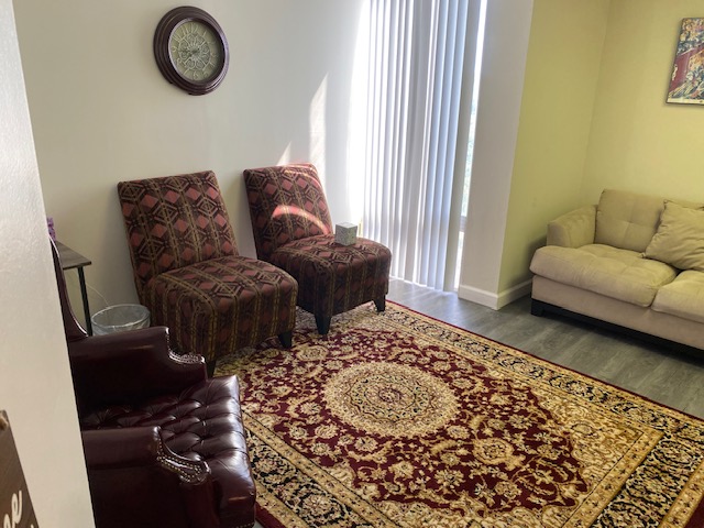 A living room with two patterned chairs, a maroon leather armchair, a beige sofa, a decorative rug, a wall clock, and sunlight streaming through vertical blinds.