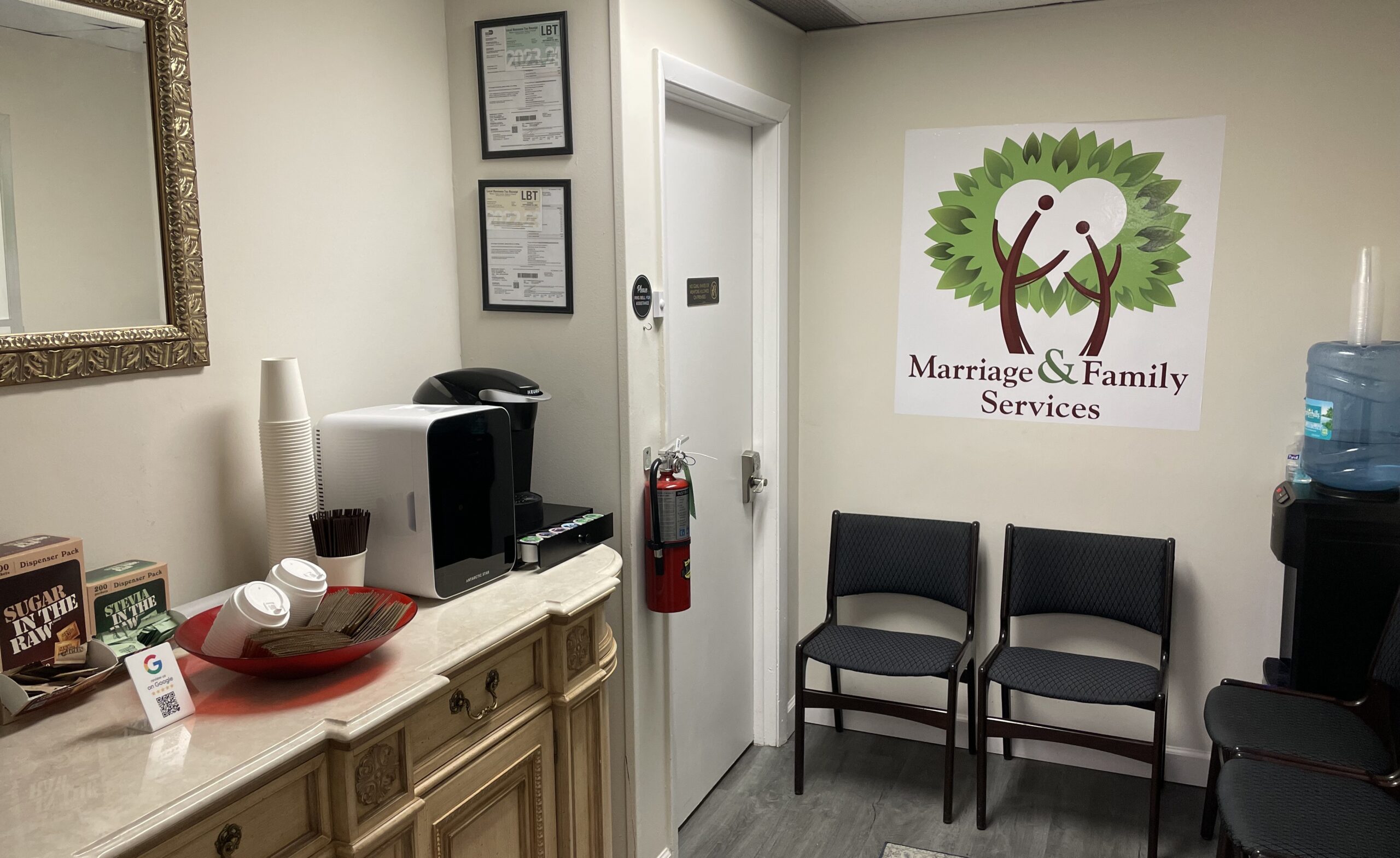 Small waiting area with chairs, a coffee station, a water cooler, and a "Marriage & Family Services" logo on the wall. Door and framed certificates are also visible.