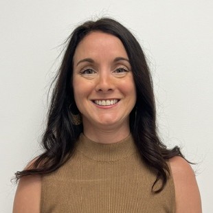 A woman with long dark hair wearing a sleeveless brown top smiles at the camera against a plain white background.