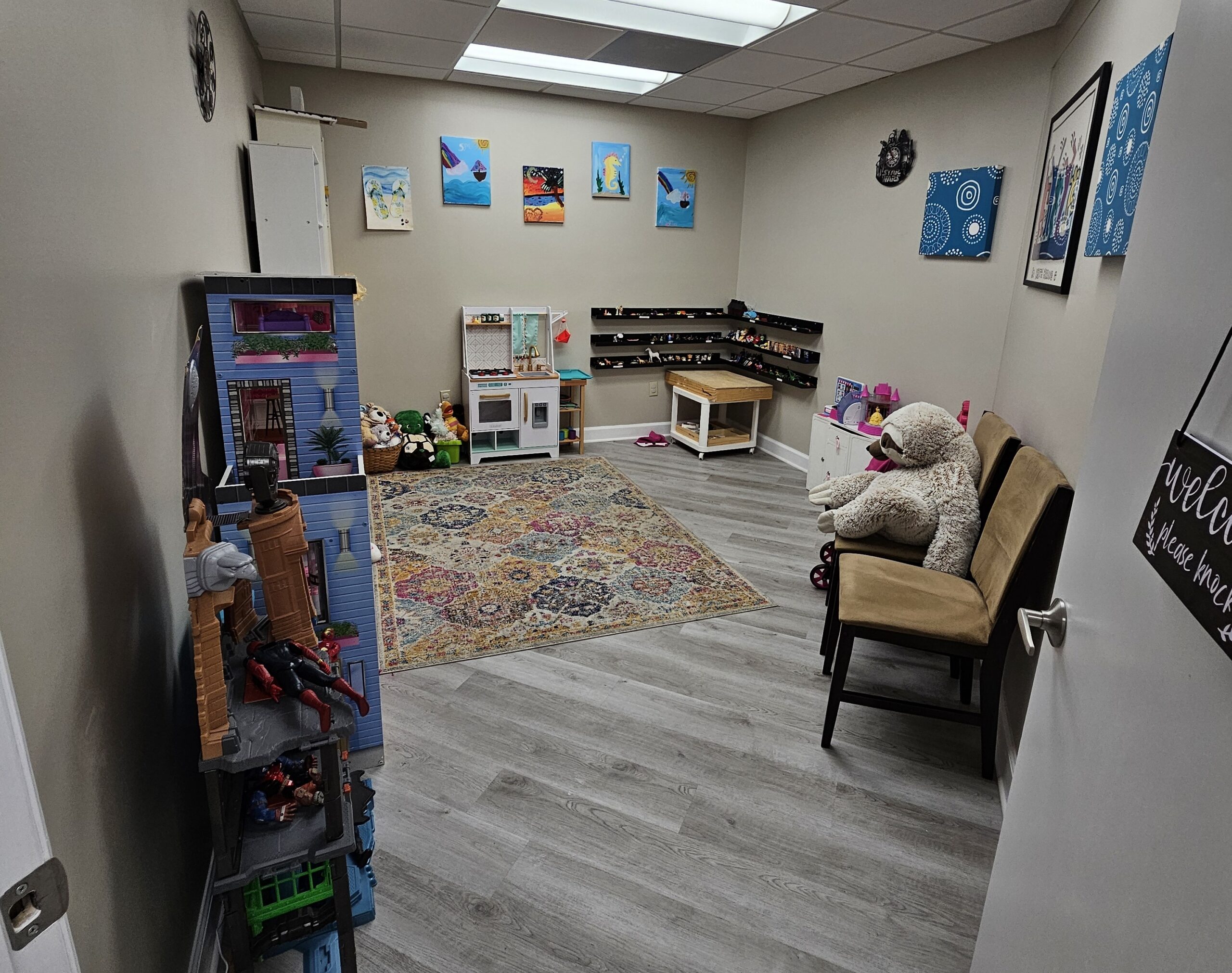 A playroom with toys, a dollhouse, play kitchen, chairs, wall art, and a large rug on a gray wooden floor. Stuffed animals and various toys are arranged around the room.