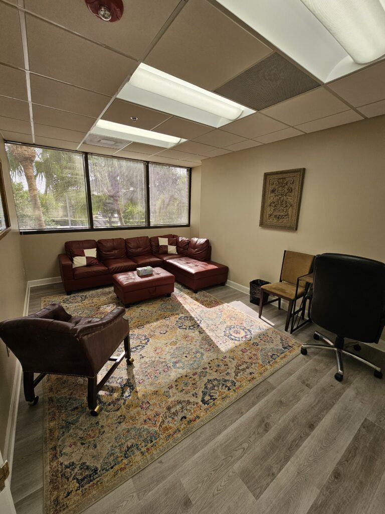 A small office with a red sectional sofa, armchair, rug, desk, black chair, and large windows letting in natural light.
