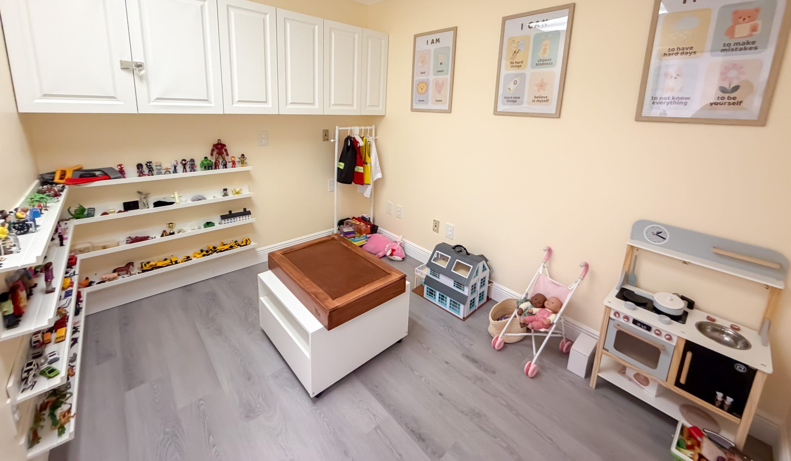 A tidy playroom with shelves of toys, a table in the center, toy kitchen, dollhouse, dress-up rack, stroller, and posters on a light yellow wall.