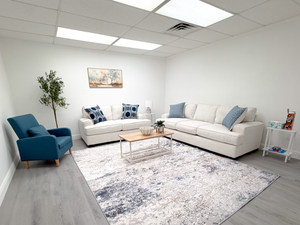 A modern living room with white sofas, a blue armchair, a patterned rug, a coffee table, a small indoor tree, and wall art in a bright, minimalist setting.