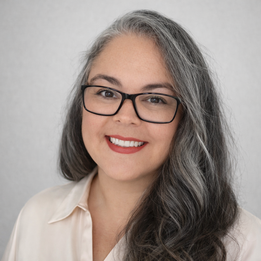 A woman with long, wavy gray hair and glasses smiles at the camera, wearing a light-colored blouse against a plain background.