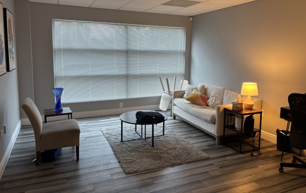 Modern living room with a gray chair, white sofa, circular coffee table, shag rug, and side tables. A large window with blinds lets in natural light. Some chairs are stacked on the sofa.