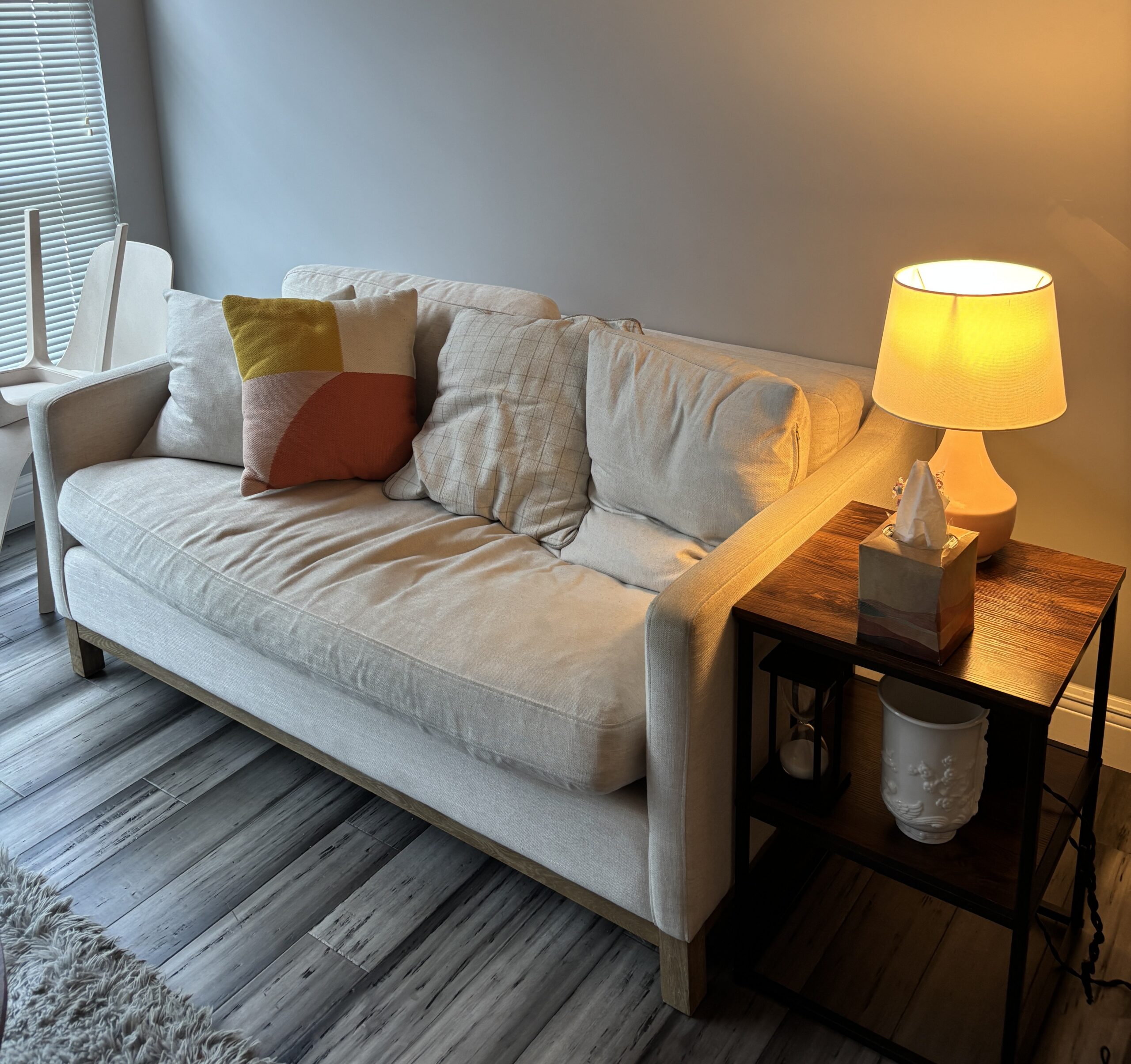 A beige sofa with two decorative pillows next to a wooden side table holding a lamp and a tissue box in a living room with gray walls and wood flooring.