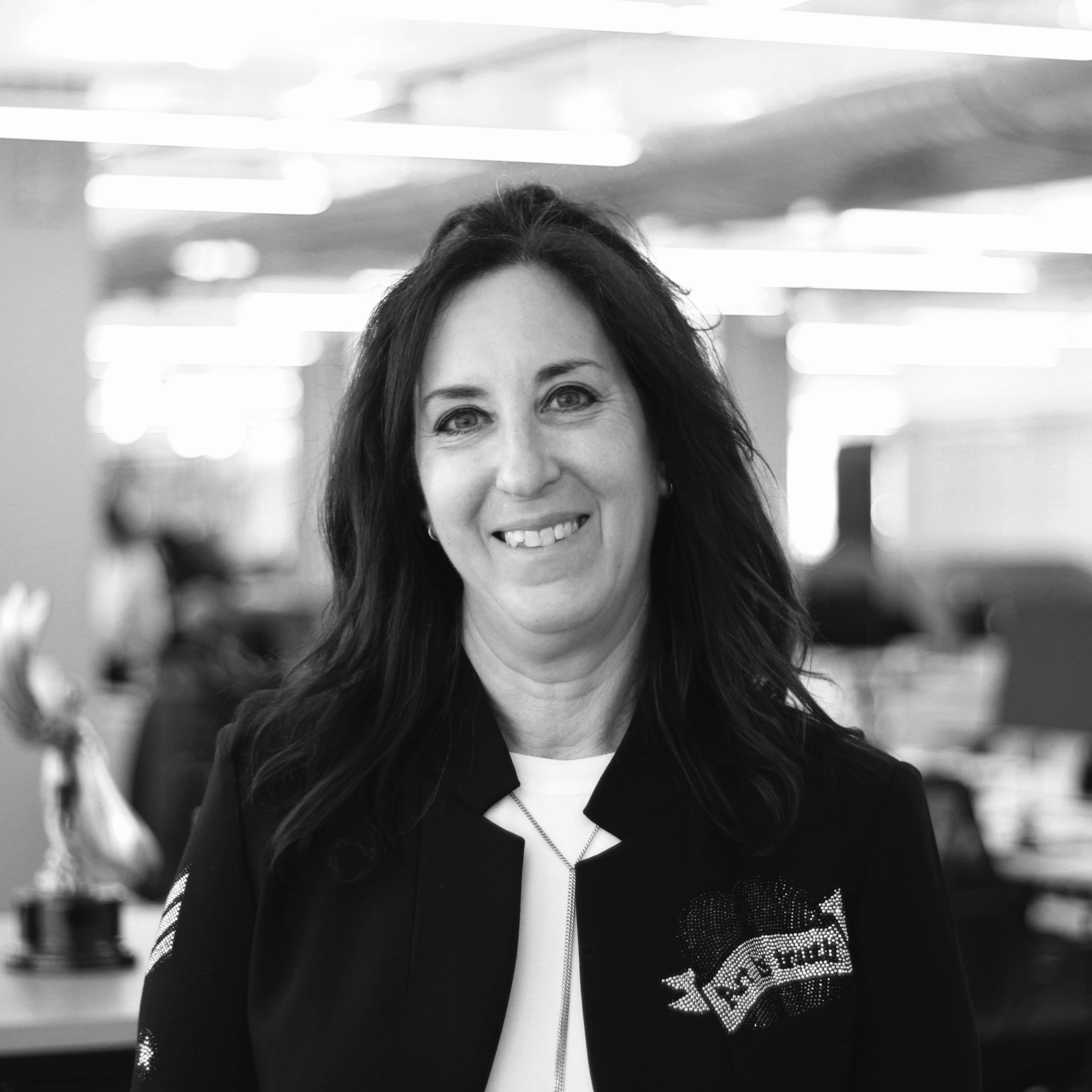 A woman with long dark hair smiles at the camera in an office setting with blurred desks and lights in the background.