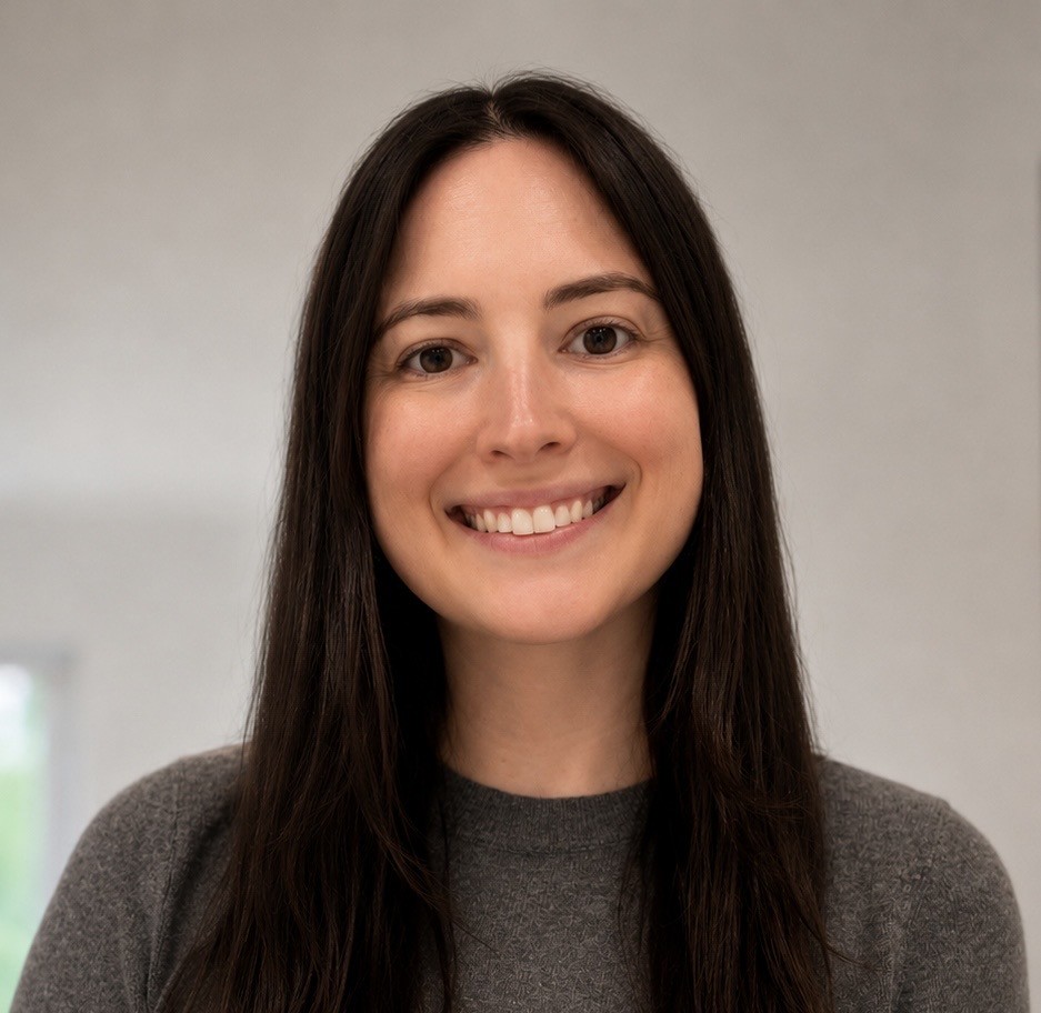 A woman with long dark hair and a grey sweater smiles at the camera against a plain, light-colored background.