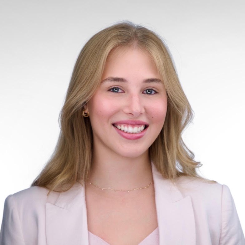 A young woman with long blonde hair, wearing a light pink blazer and top, smiles at the camera against a plain light background.
