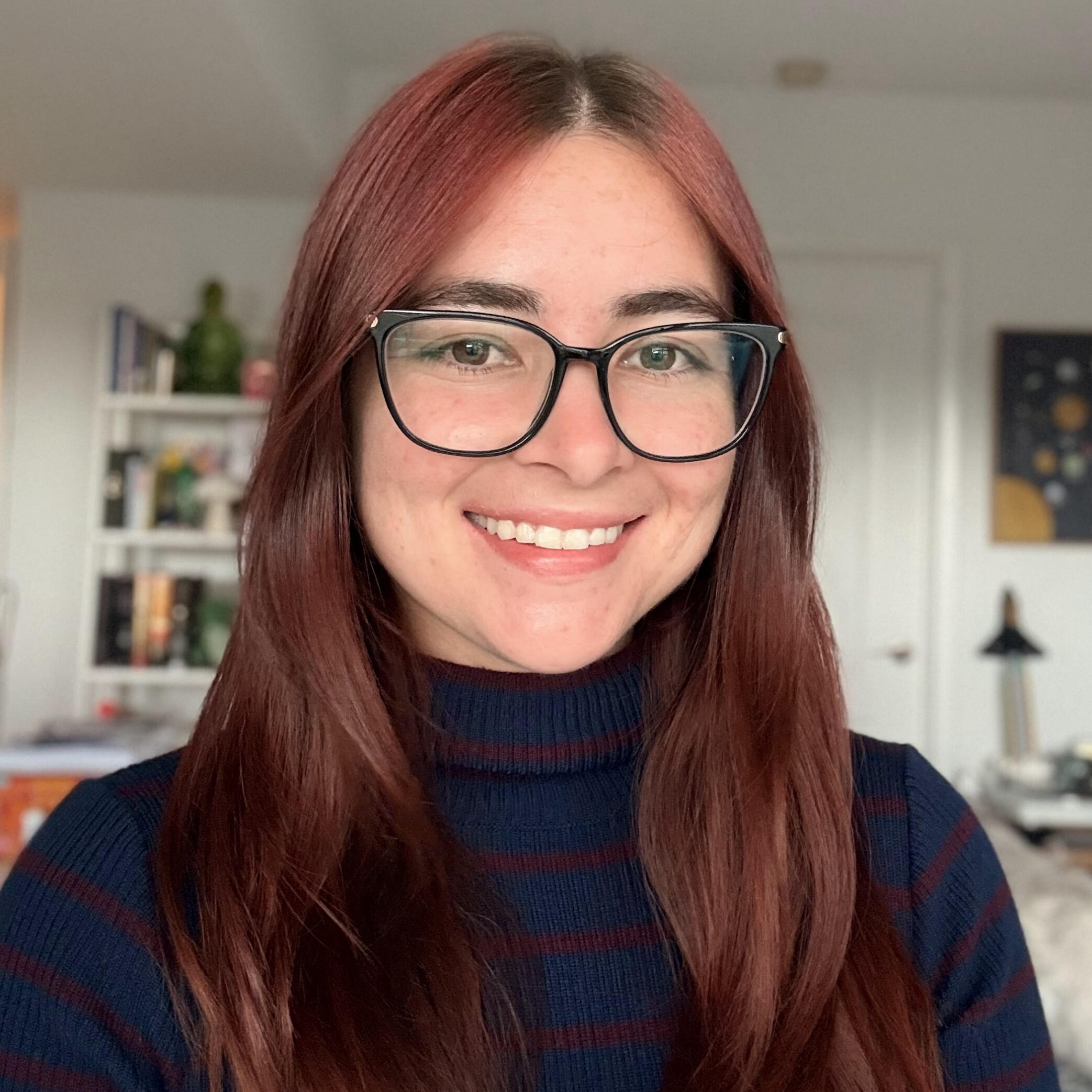 A person with long, reddish-brown hair and glasses smiles at the camera, wearing a dark striped turtleneck in a well-lit room with shelves and decor in the background.