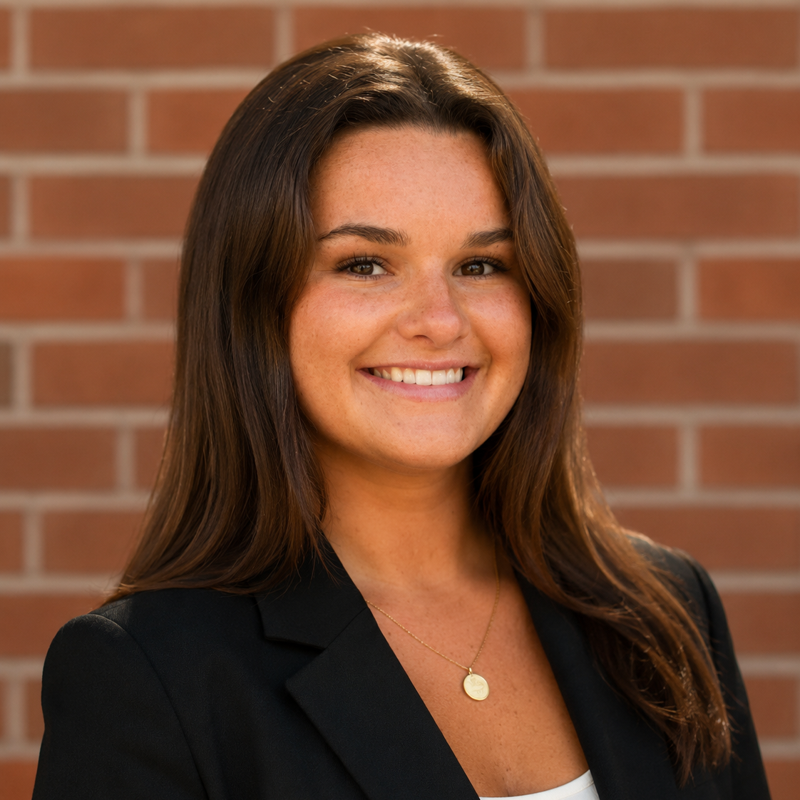 Woman with long brown hair wearing a black blazer and white top, smiling in front of a red brick wall.