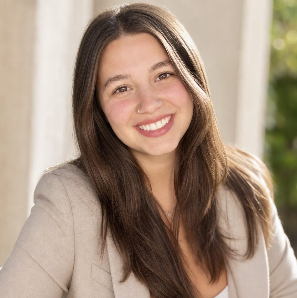 A woman with long brown hair and a beige blazer smiles at the camera, standing outdoors in natural light.