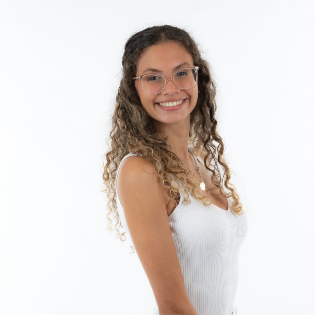 Smiling young woman with curly hair and glasses, wearing a white sleeveless top, standing against a plain white background.