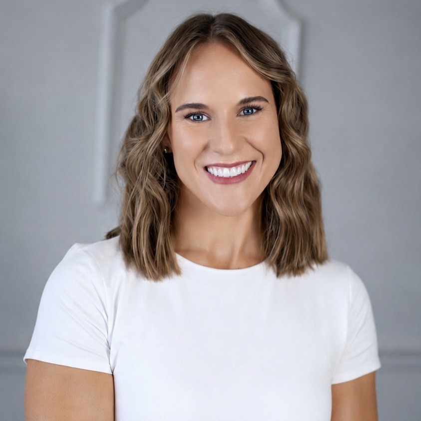 Woman with wavy light brown hair and a white shirt smiles at the camera against a gray background.