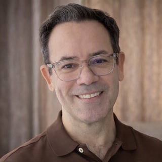 A man with short dark hair and glasses smiles at the camera, wearing a brown polo shirt with a blurred brown background.
