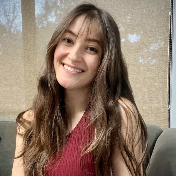 A woman with long brown hair, wearing a sleeveless burgundy top, smiles while sitting on a gray couch in front of a window with beige blinds.