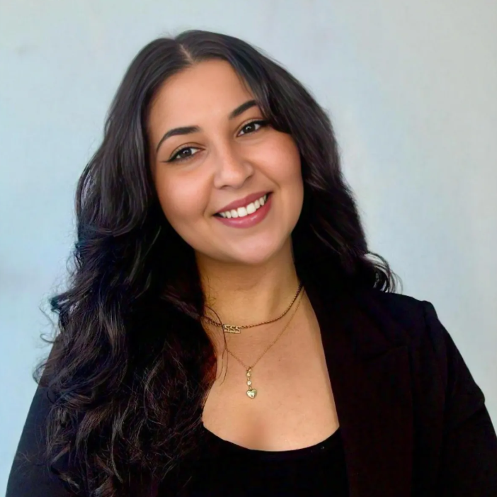 Woman with long dark hair wearing a black top and blazer, smiling at the camera against a plain light background.