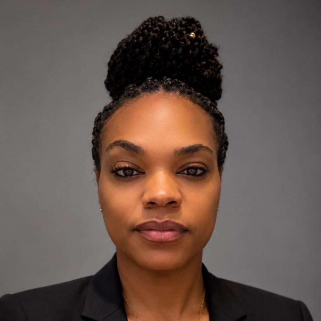A woman with braided hair in a bun, wearing a black blazer and a gold necklace, poses in front of a plain gray background.