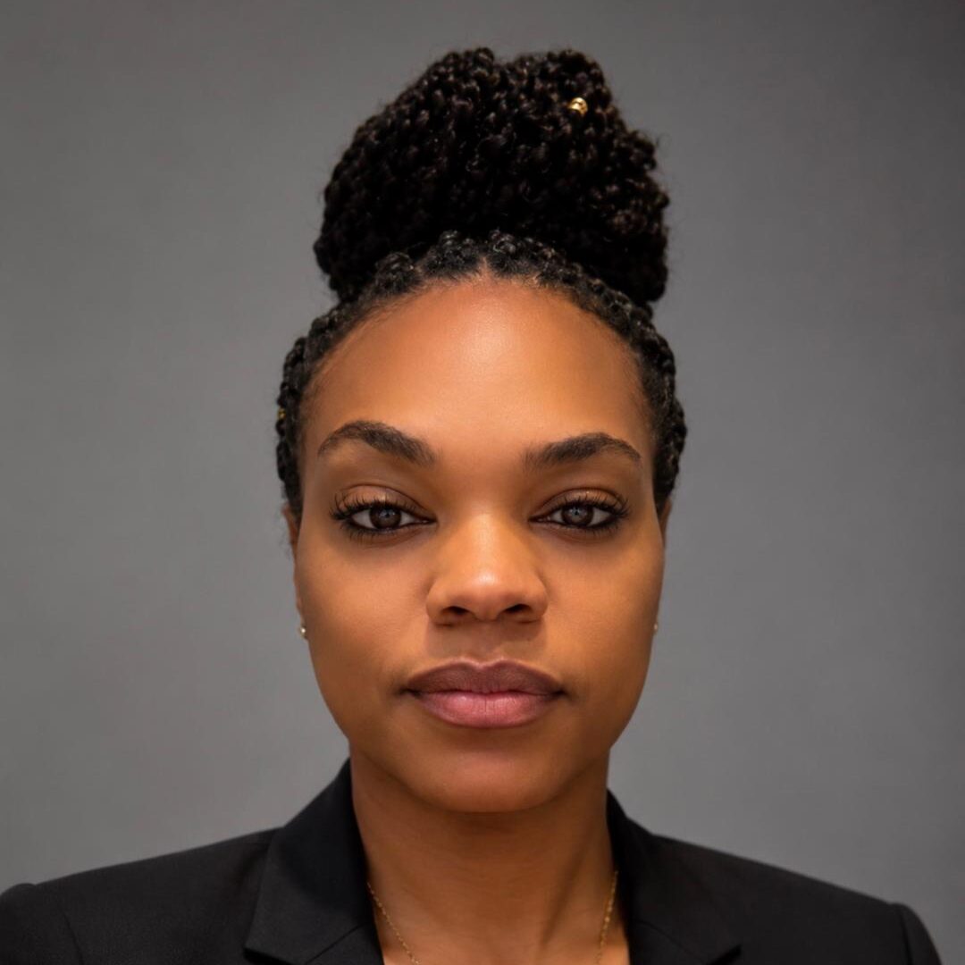 A woman with braided hair in a bun, wearing a black blazer and a gold necklace, poses in front of a plain gray background.