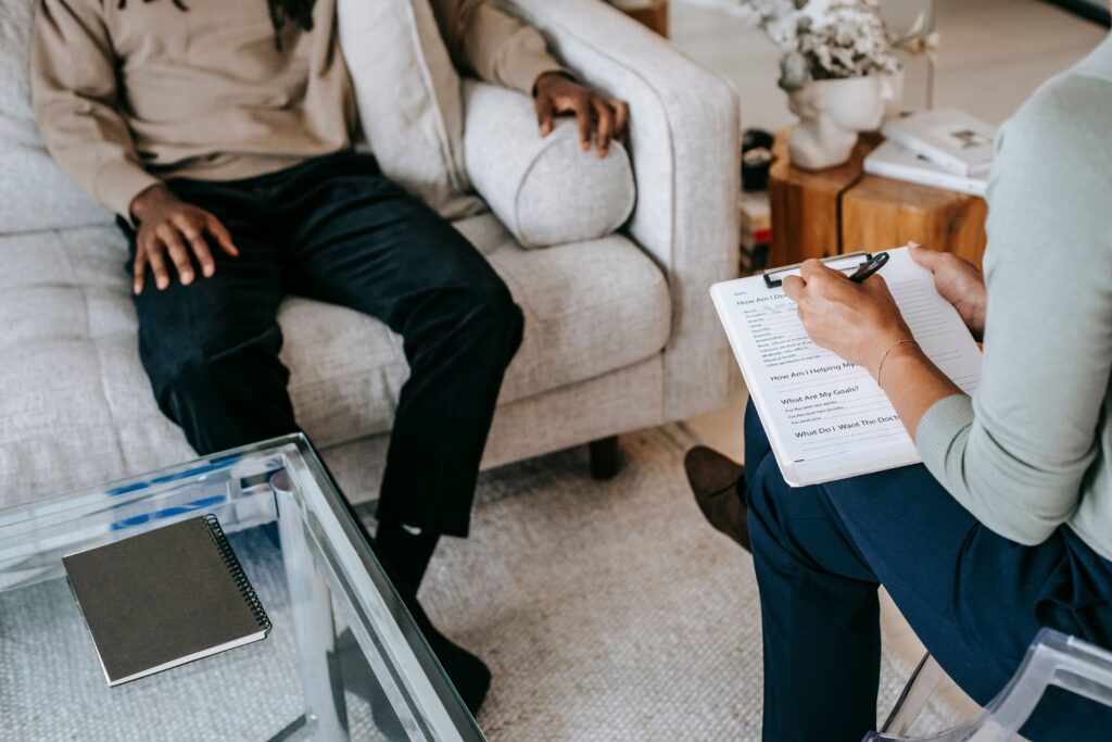 Two people sit across from each other in a living room; one person is writing on a clipboard while the other sits on a couch, partially visible. A notebook is on a glass coffee table.