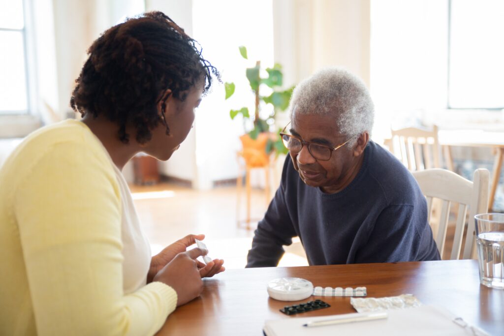 Two adults sit at a table with a pill organizer and medication, appearing to discuss or review the pills together in a well-lit room.